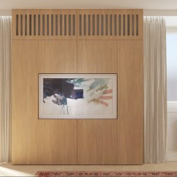 BEZPIECZNYWYKONAWCA.PL SPÓŁKA Z OGRANICZONĄ ODPOWIEDZIALNOŚCIĄ - Drewniana zabudowa ściany z ukrytym telewizorem i szafkami, jasne zasłony, okna z widokiem na zieleń, grzejnik żeliwny, dywanik w orientalny wzór.