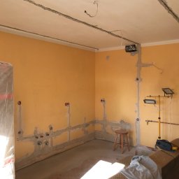 BEZPIECZNYWYKONAWCA.PL SPÓŁKA Z OGRANICZONĄ ODPOWIEDZIALNOŚCIĄ - Żółte ściany w trakcie remontu, widoczne instalacje elektryczne i hydrauliczne, przygotowane do dalszych prac wykończeniowych. Stojąca lampa budowlana oświetla wnętrze.