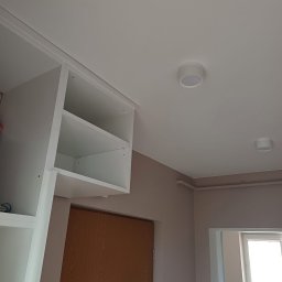 BEZPIECZNYWYKONAWCA.PL SPÓŁKA Z OGRANICZONĄ ODPOWIEDZIALNOŚCIĄ - Białe, nowoczesne półki nad drzwiami wejściowymi, z widocznymi dwoma okrągłymi lampami sufitowymi. Ujęcie z dołu, podkreślające minimalistyczny design i jasne kolory.