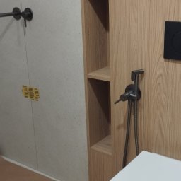 BEZPIECZNYWYKONAWCA.PL SPÓŁKA Z OGRANICZONĄ ODPOWIEDZIALNOŚCIĄ - Nowoczesna łazienka po remoncie: biała wisząca toaleta z bidetką, drewniane półki w zabudowie, szare płytki. Minimalistyczny design z akcentami w kolorze drewna.