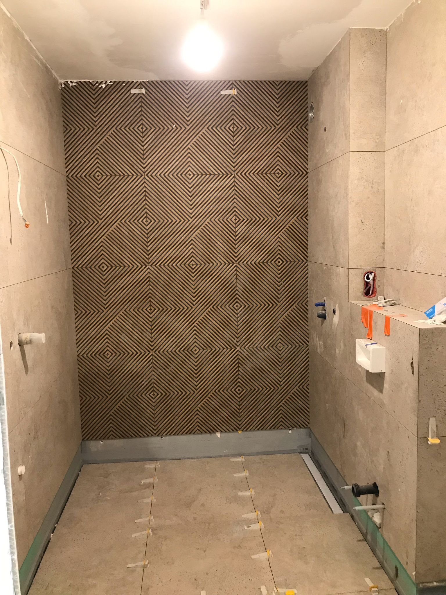 Wnętrze łazienki w trakcie wykańczania. Na ścianach beżowe płytki, jedna ściana z dekoracyjnym wzorem. Na podłodze płytki z krzyżykami dystansowymi.