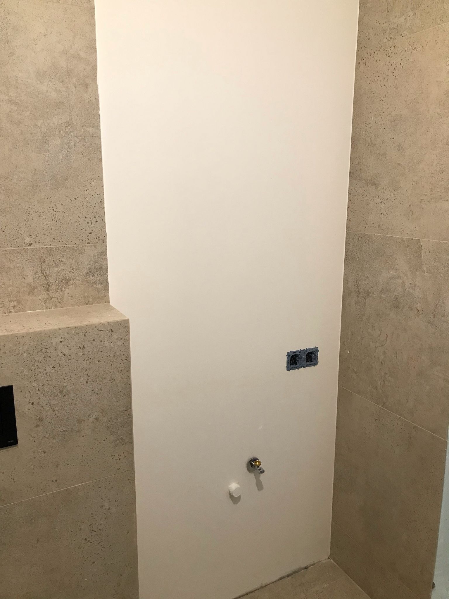 Biała ściana z gładzią, widoczne gniazdka elektryczne i przyłącze wodne w łazience wykończonej płytkami w odcieniach beżu. Minimalistyczny design.