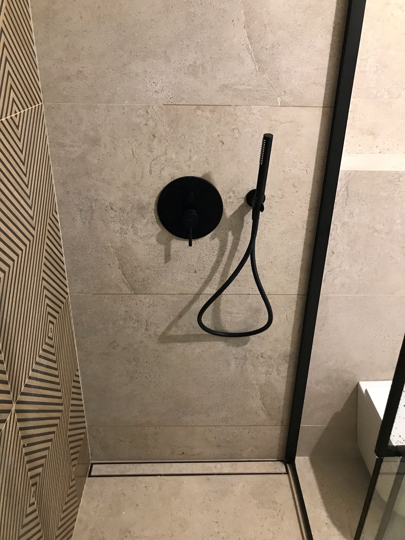 Nowoczesna kabina prysznicowa z czarną armaturą i geometryczną mozaiką. Beżowe płytki imitujące kamień tworzą minimalistyczne tło.