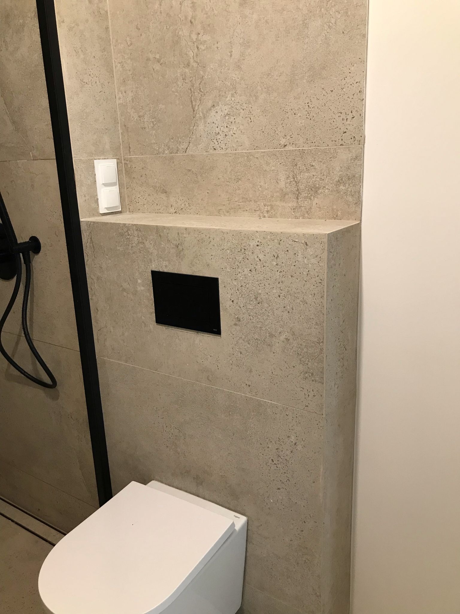 Nowoczesna toaleta z beżowymi płytkami imitującymi kamień, czarnym panelem sterowania i białą miską WC. Minimalistyczny design, eleganckie wykończenie.