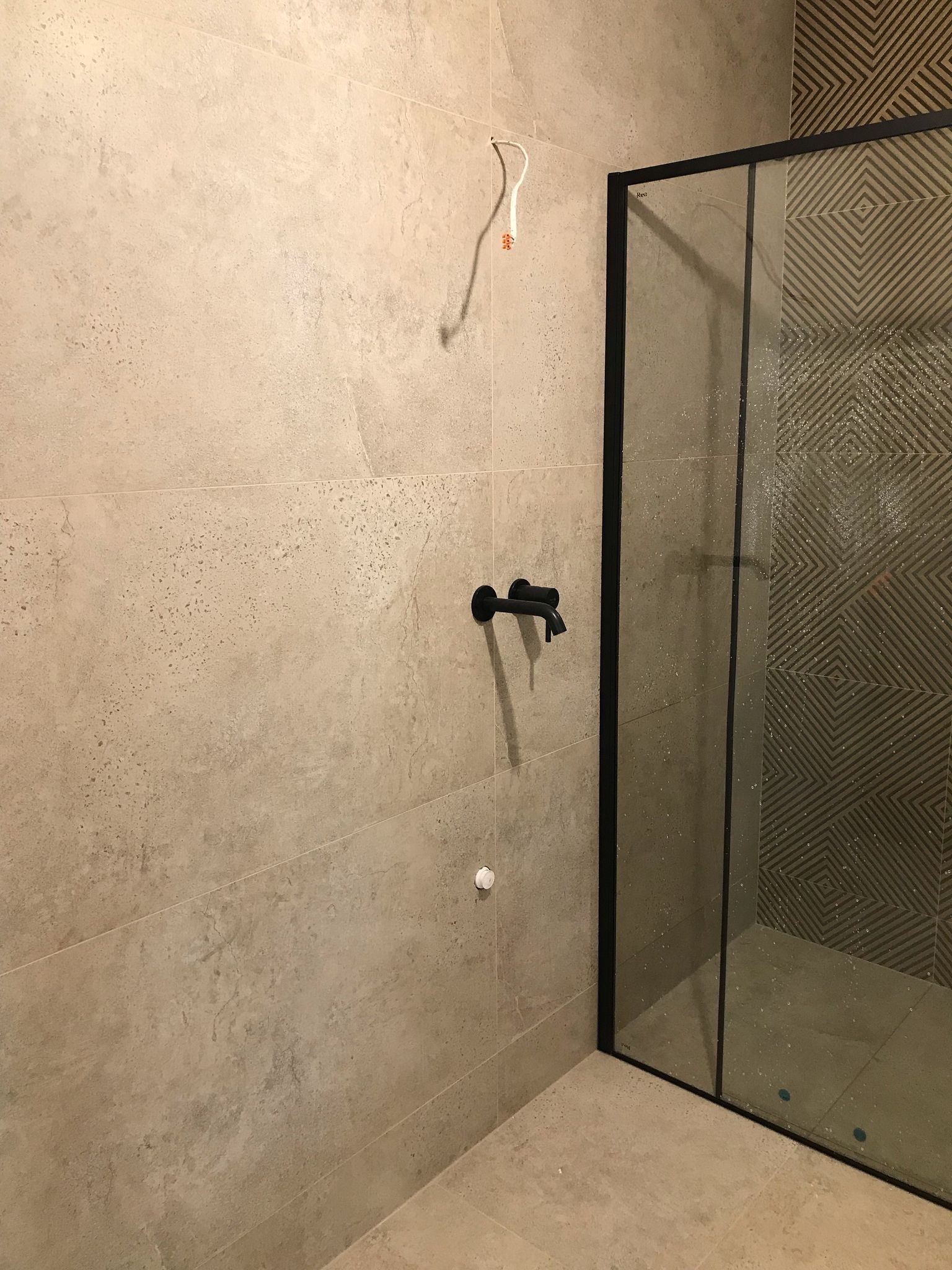 Wykończona łazienka z beżowymi płytkami, czarną armaturą i kabiną prysznicową z geometrycznym wzorem na ścianie. Widoczne wystające przewody elektryczne.