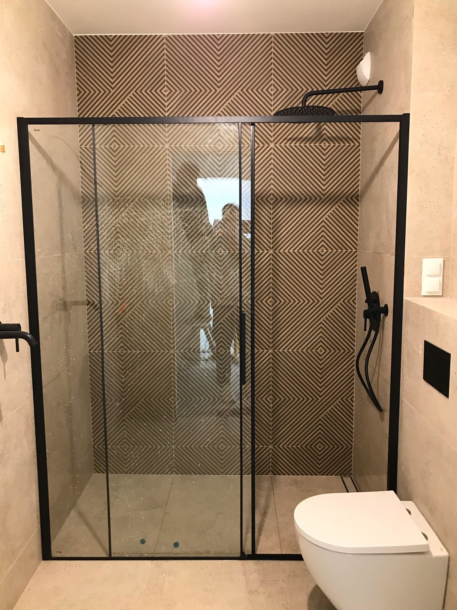 Nowoczesna kabina prysznicowa z czarnymi ramami i deszczownicą na tle płytek z geometrycznym wzorem. Widoczna toaleta podwieszana i armatura w czarnym macie.