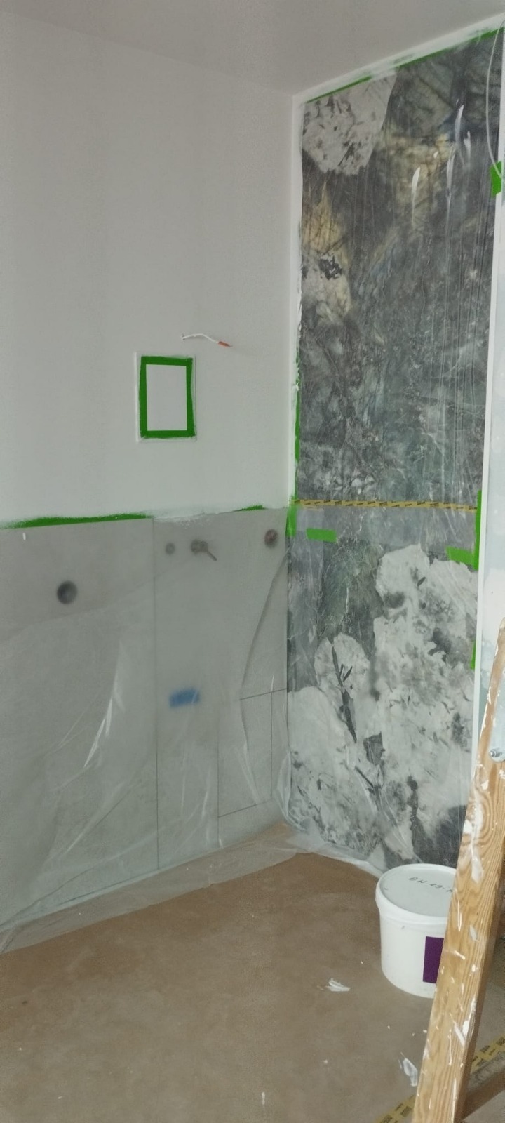 Łazienka w trakcie prac: zabezpieczone folią płytki i ściana z efektem marmuru, widoczna drabina i wiadro. Przygotowania do dalszych prac wykończeniowych.