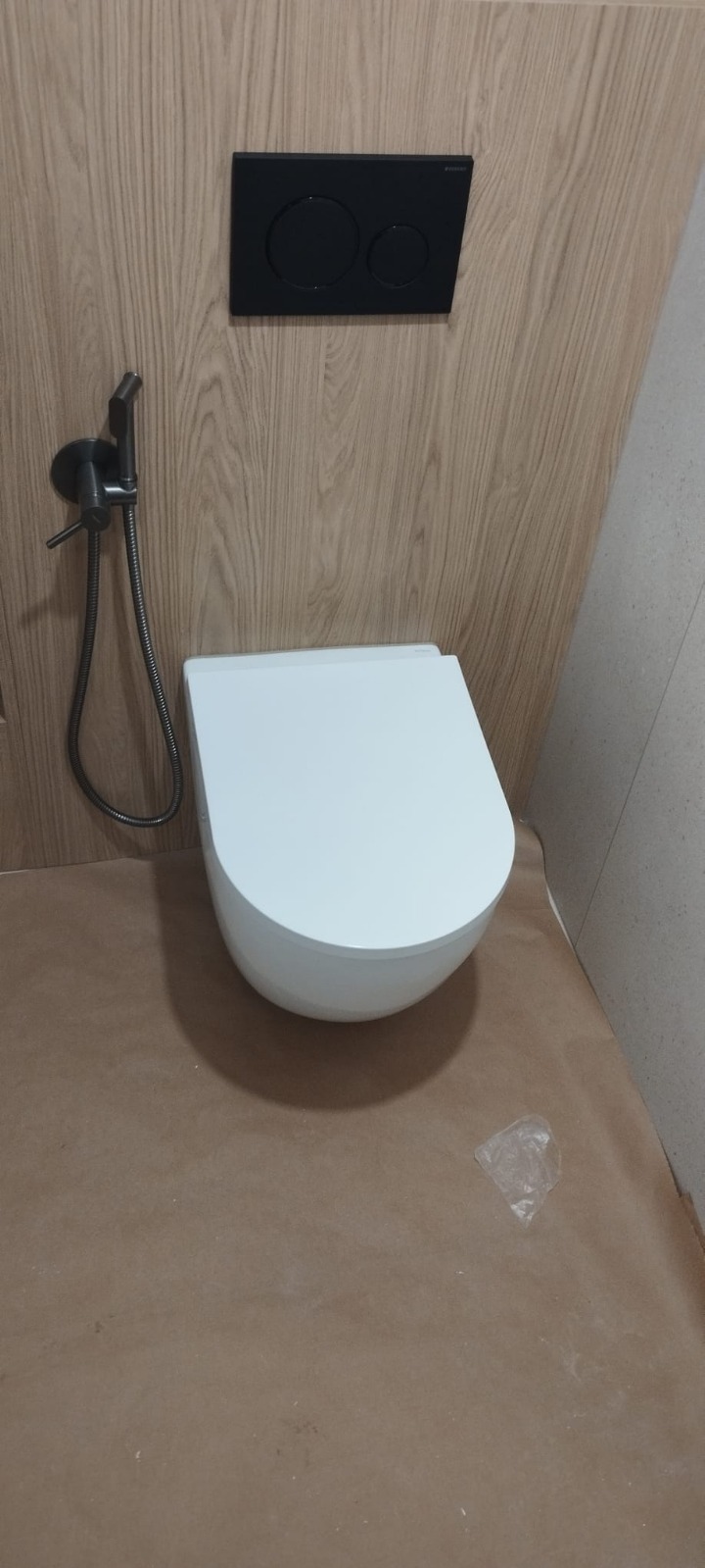 Biała, podwieszana miska WC z czarnym przyciskiem spłukującym i bidetką na tle drewnopodobnej okładziny ściennej. Podłoga zabezpieczona kartonem.