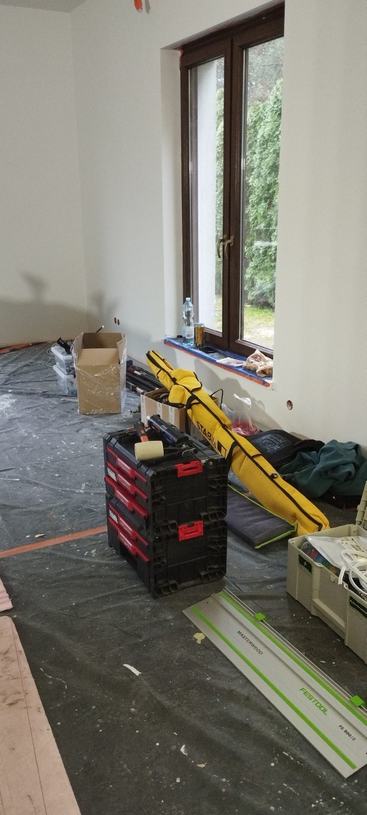 Przygotowania do prac wykończeniowych w Warszawie: zabezpieczona folią podłoga, narzędzia glazurnicze, skrzynki narzędziowe. Świeżo pomalowane ściany, okno.