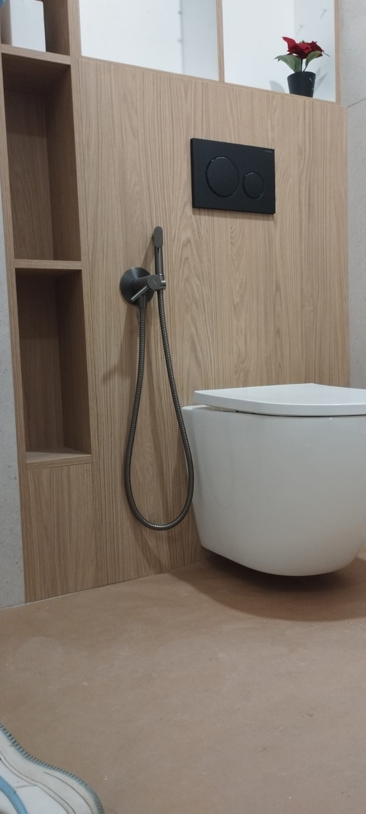 Nowoczesna toaleta podwieszana z drewnianą zabudową, czarnym przyciskiem spłukującym i bidetką w łazience. Minimalistyczny design z półkami i rośliną.