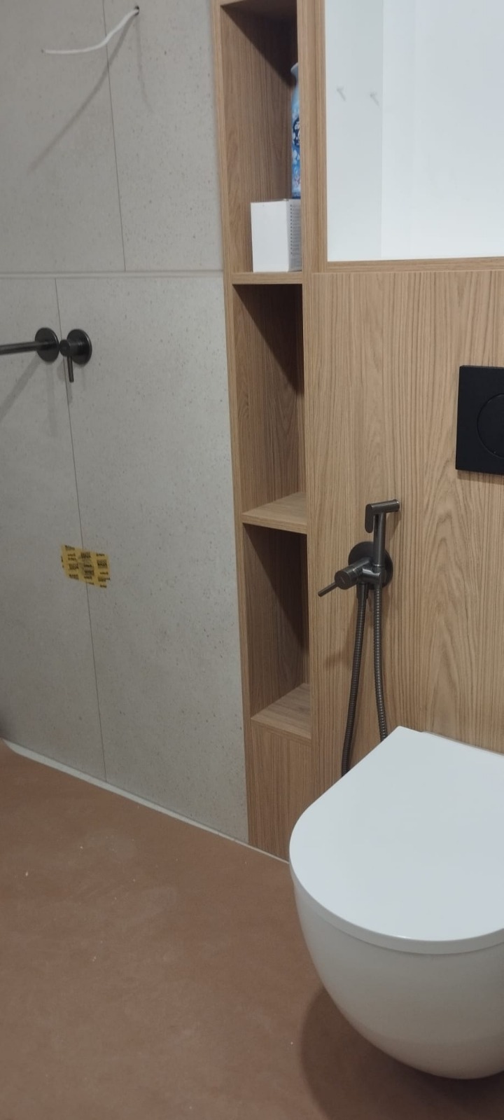 Nowoczesna łazienka po remoncie: biała wisząca toaleta z bidetką, drewniane półki w zabudowie, szare płytki. Minimalistyczny design z akcentami w kolorze drewna.