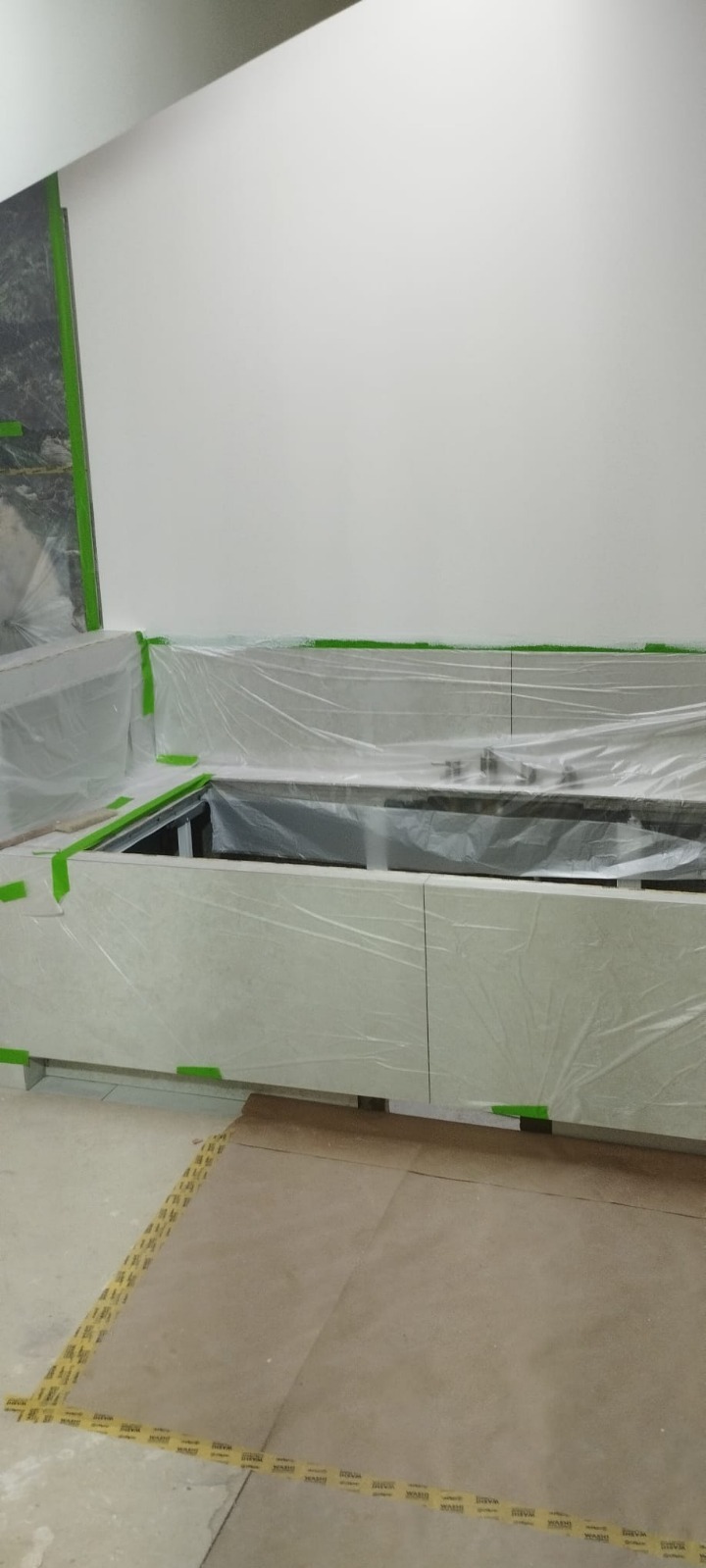 Przygotowanie do prac wykończeniowych: zabezpieczona folią i taśmą maskującą zabudowa kuchenna z jasnego kamienia, ściana przygotowana do malowania.