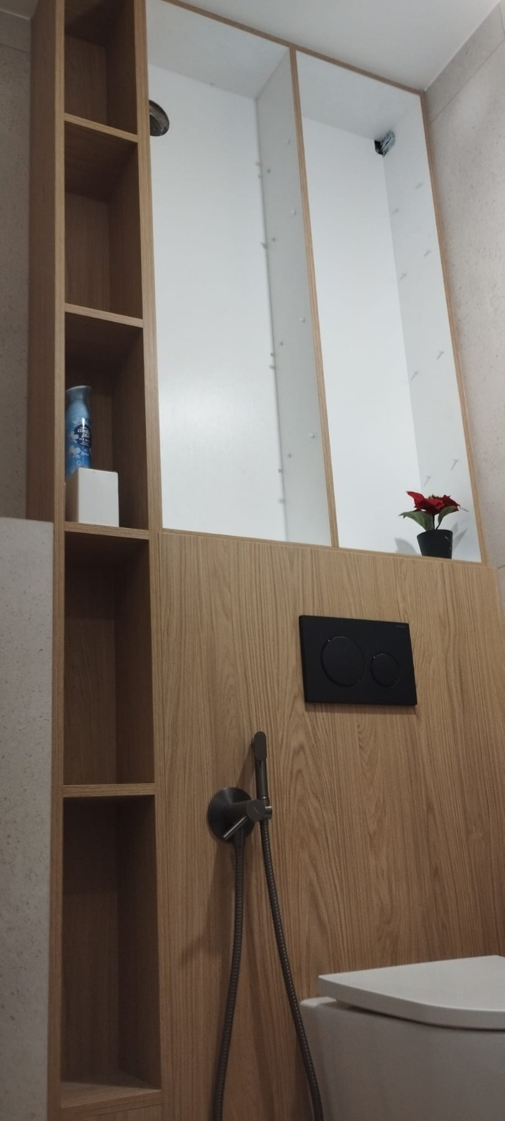 Nowoczesna łazienka z zabudową WC w odcieniach drewna, z półkami i ukrytą spłuczką. Minimalistyczny design z akcentem roślinnym i armaturą w kolorze grafitu.