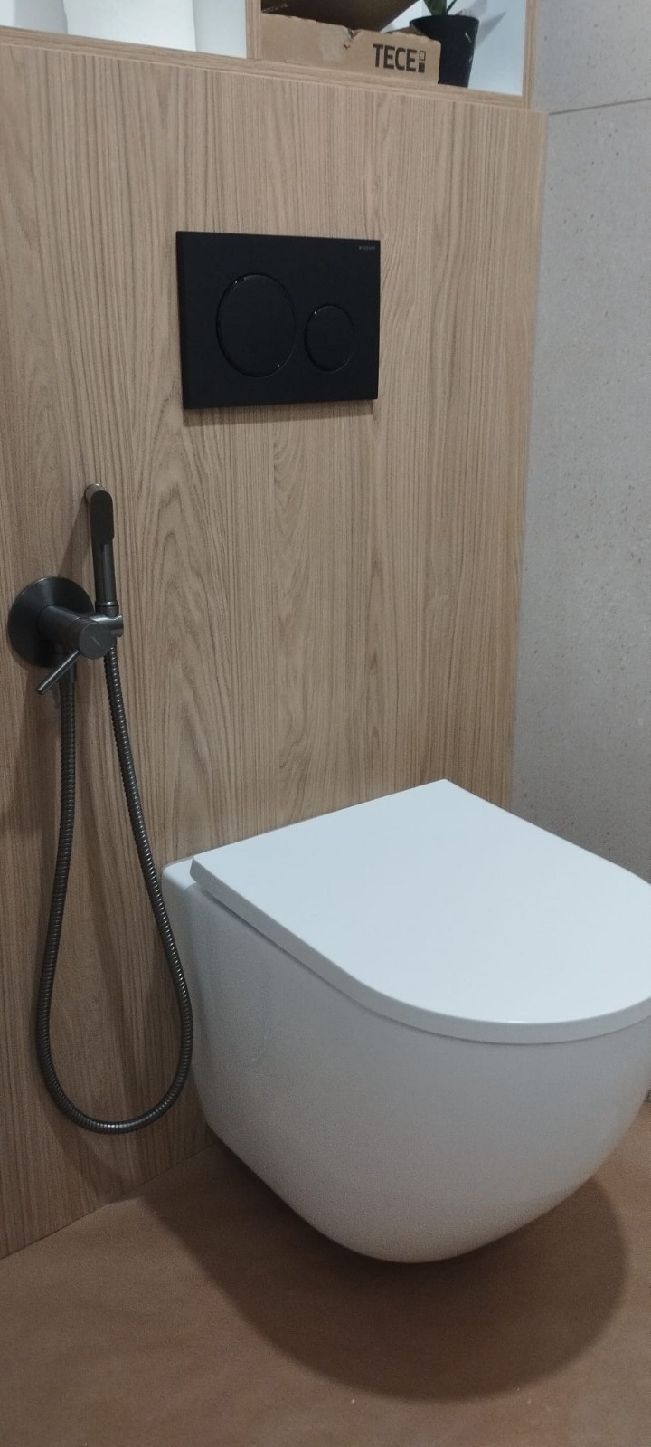 Nowoczesna toaleta podwieszana z bidetką i czarnym panelem sterującym TECE na drewnopodobnej ściance. Minimalistyczny design, wykończenie łazienki.