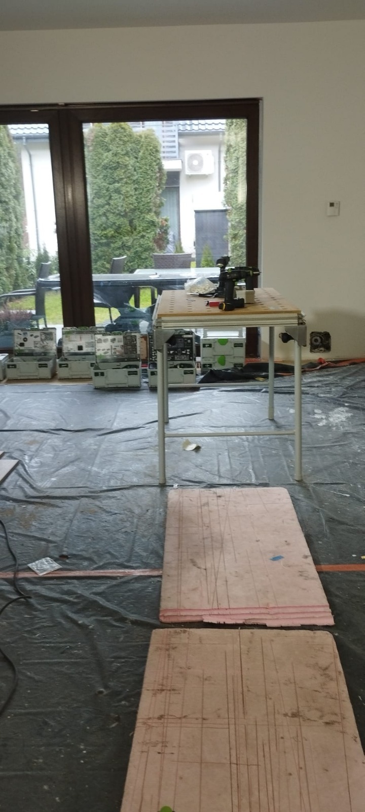 Przygotowania do prac wykończeniowych: stół z narzędziami, skrzynki narzędziowe i płyty podłogowe z naniesionymi liniami na folii ochronnej w jasnym wnętrzu.