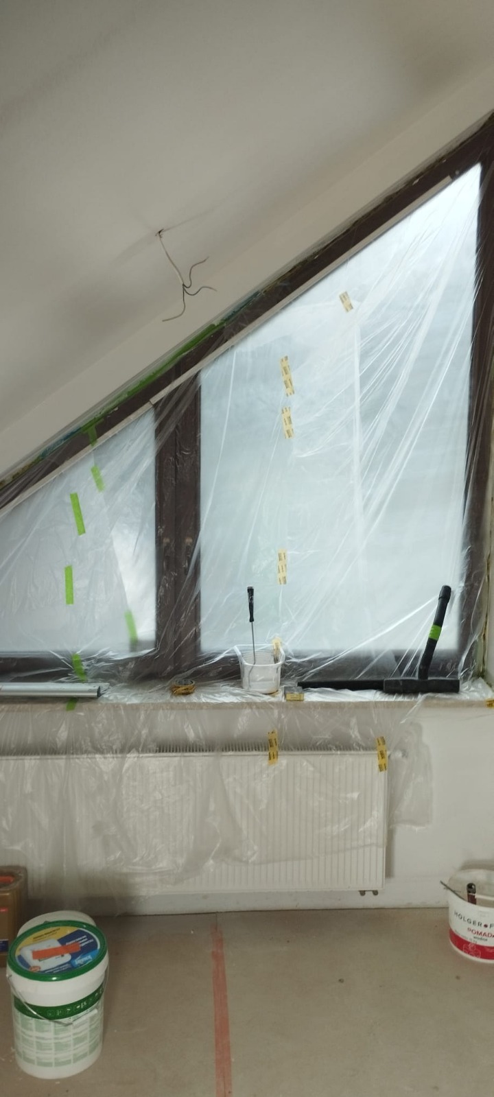 Skośne okno zabezpieczone folią malarską, grzejnik osłonięty, widoczne narzędzia malarskie i farby. Przygotowania do malowania w pokoju na poddaszu.