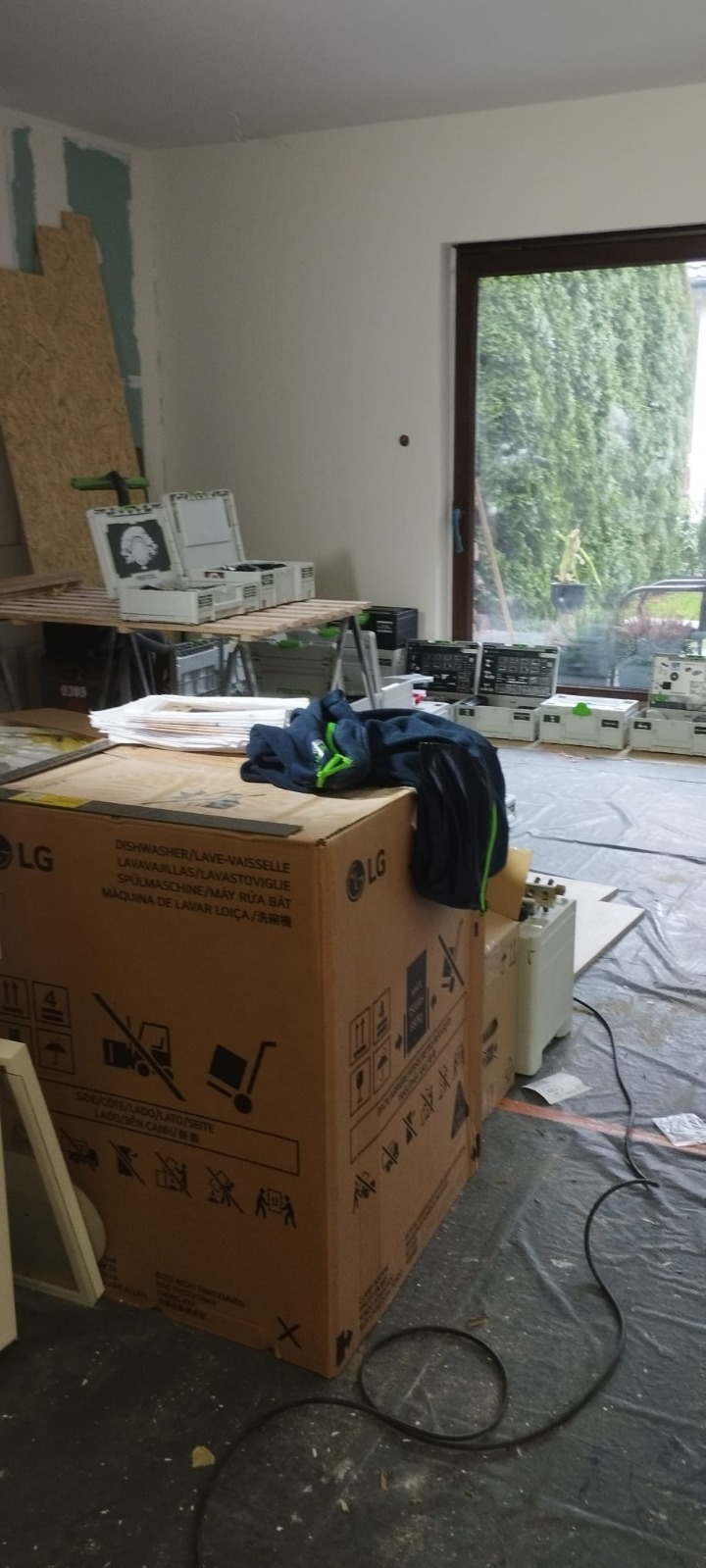 Wnętrze w trakcie prac wykończeniowych: karton LG, narzędzia Festool, zabezpieczona folią podłoga. Widoczne ślady szpachlowania na ścianach. Okno z widokiem na zieleń.