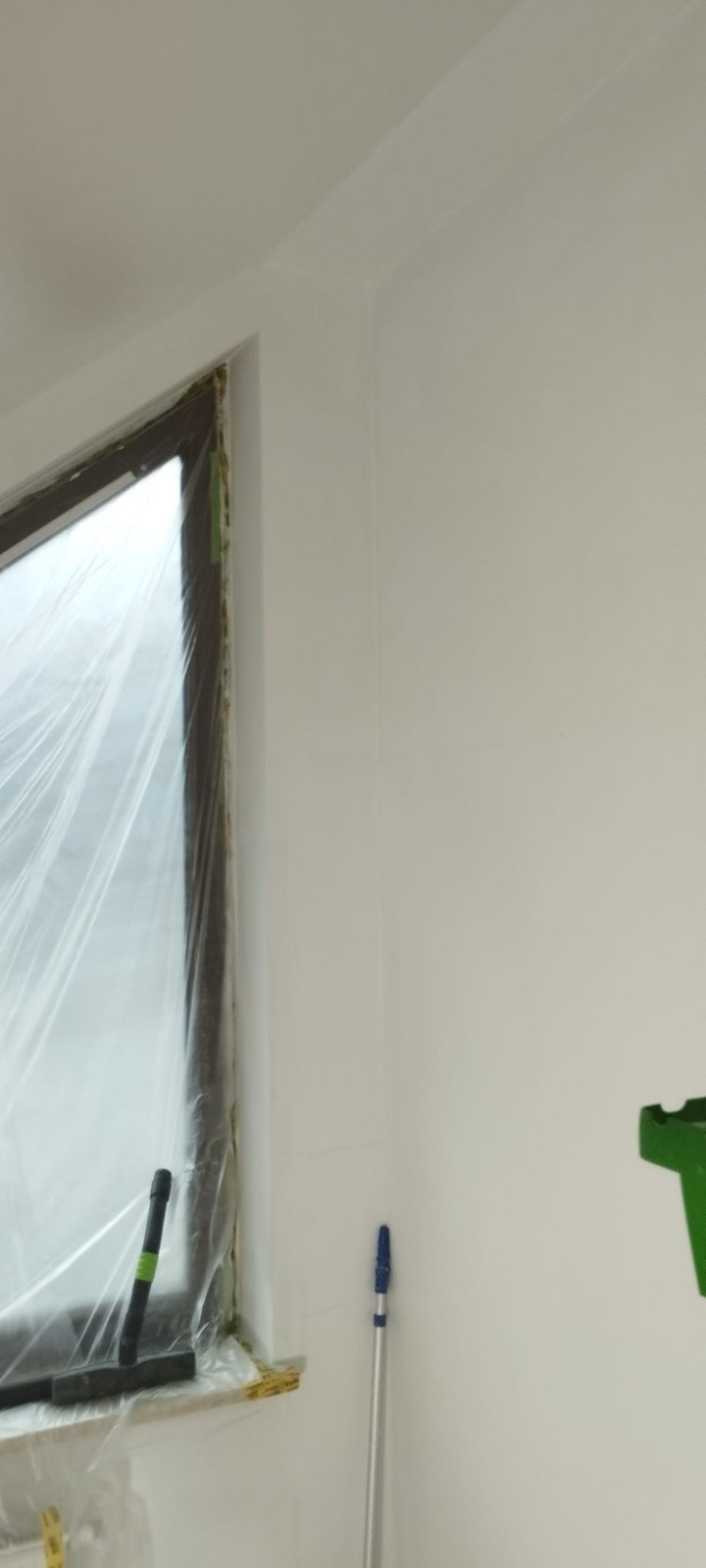 Biała ściana po malowaniu, okno zabezpieczone folią malarską, widoczny wałek i kuweta malarska. Świeżo pomalowane narożniki i listwy przysufitowe.