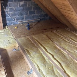 Usługi budowlane KF - Ocieplenie poddasza wełną mineralną, widoczne drewniane belki i płyty OSB. Na płycie wkrętarka i miarka. Fragment ściany z bloczków.