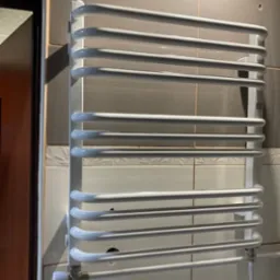 Biały, drabinkowy grzejnik łazienkowy zamontowany na kafelkowanej ścianie. Widoczne przyłącza hydrauliczne i zawór termostatyczny.