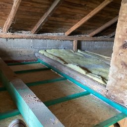 Usługi budowlane KF - Poddasze w trakcie ocieplania: zielone belki konstrukcyjne, płyty OSB i fragment wełny mineralnej. Widoczny fragment buta. Drewniana więźba dachowa.