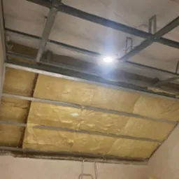 Montaż izolacji z wełny mineralnej w konstrukcji sufitu podwieszanego z profili metalowych. Widoczne oświetlenie robocze.