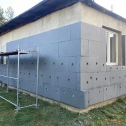 Ocieplanie ściany budynku szarym styropianem, widoczne rusztowanie. Częściowo ocieplona ściana z widocznymi otworami montażowymi.