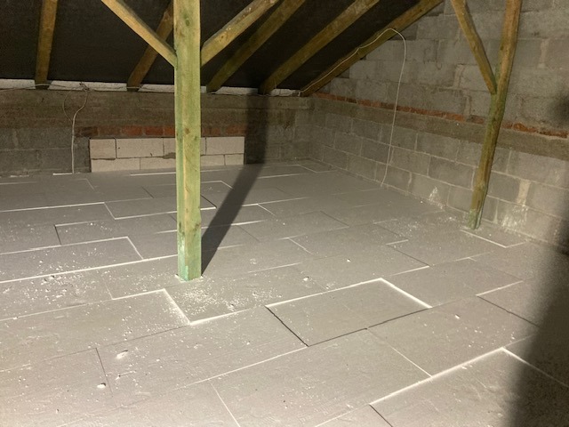 Szare płyty izolacyjne ułożone na podłodze poddasza, widoczne drewniane elementy konstrukcji dachu i słup podtrzymujący. Prace wykończeniowe w surowym wnętrzu.