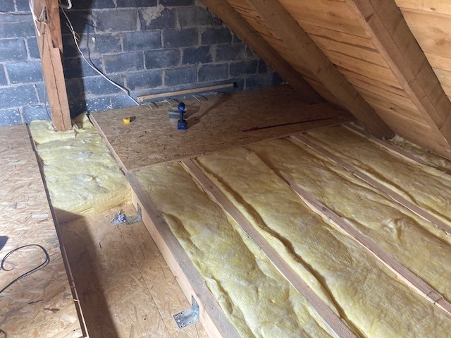 Ocieplenie poddasza wełną mineralną, widoczne drewniane belki i płyty OSB. Na płycie wkrętarka i miarka. Fragment ściany z bloczków.