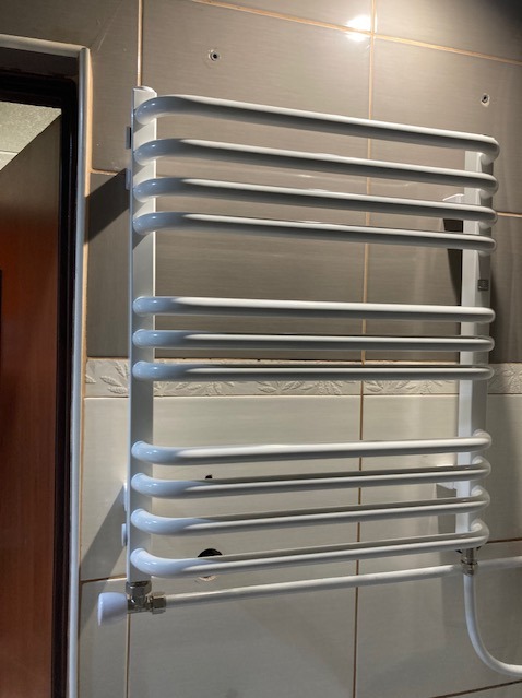 Biały, drabinkowy grzejnik łazienkowy zamontowany na kafelkowanej ścianie. Widoczne przyłącza hydrauliczne i zawór termostatyczny.