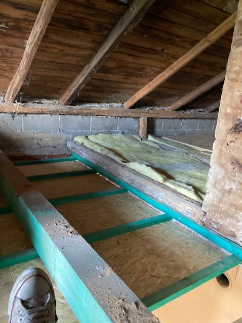 Poddasze w trakcie ocieplania: zielone belki konstrukcyjne, płyty OSB i fragment wełny mineralnej. Widoczny fragment buta. Drewniana więźba dachowa.