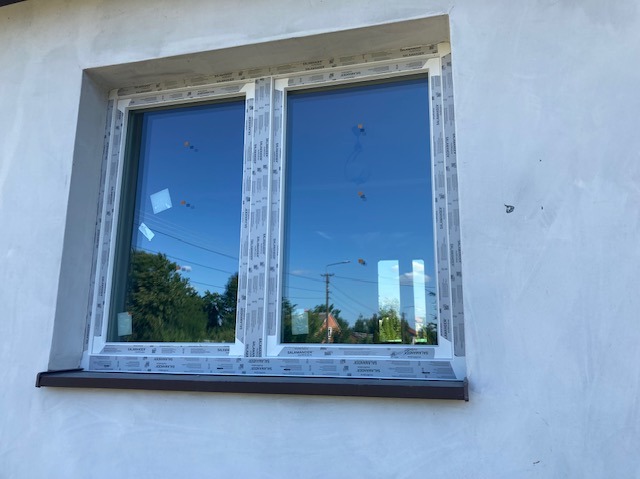 Nowe, białe okno PCV z brązowym parapetem zamontowane w surowej, jasnej elewacji. Widoczne zabezpieczenia na profilach okiennych.