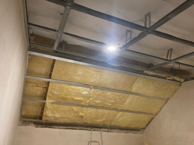 Montaż izolacji z wełny mineralnej w konstrukcji sufitu podwieszanego z profili metalowych. Widoczne oświetlenie robocze.