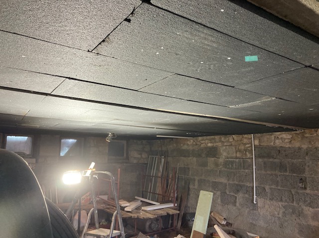 Ocieplenie sufitu w piwnicy płytami izolacyjnymi, widoczne płyty z szarego styropianu. Ściany z kamienia, okna piwniczne. Oświetlenie robocze.