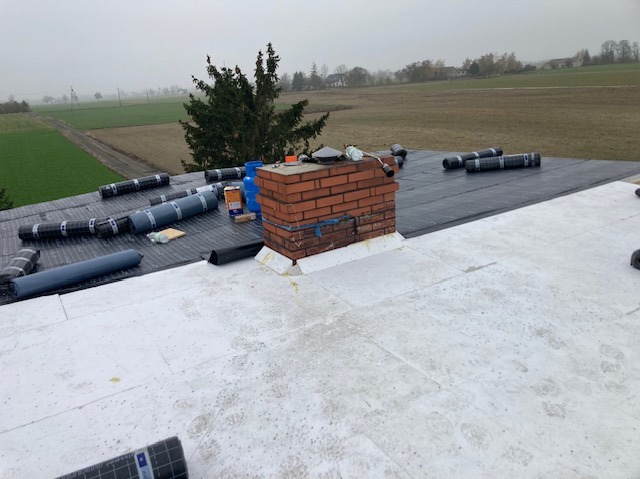 Izolacja dachu płaskiego z kominem: białe płyty izolacyjne i czarna membrana dachowa w rolkach. Widoczne narzędzia i materiały budowlane. Krajobraz w tle.