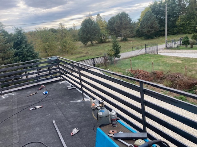 Montaż metalowej balustrady na dachu budynku z widokiem na zieleń. Widoczne narzędzia pracy: spawarka, butla z gazem, szlifierka. Prace spawalnicze w toku.