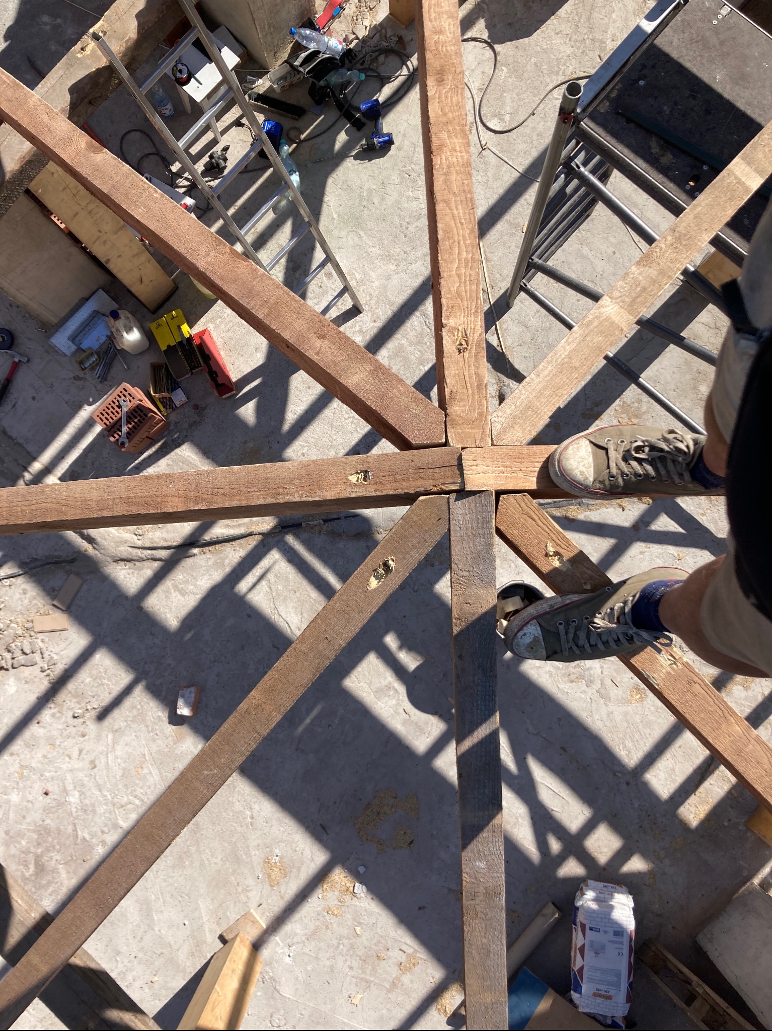 Drewniana konstrukcja dachu w trakcie budowy, widok z góry. Elementy połączone, widoczne narzędzia i drabina. Cień tworzy geometryczne wzory na podłożu.