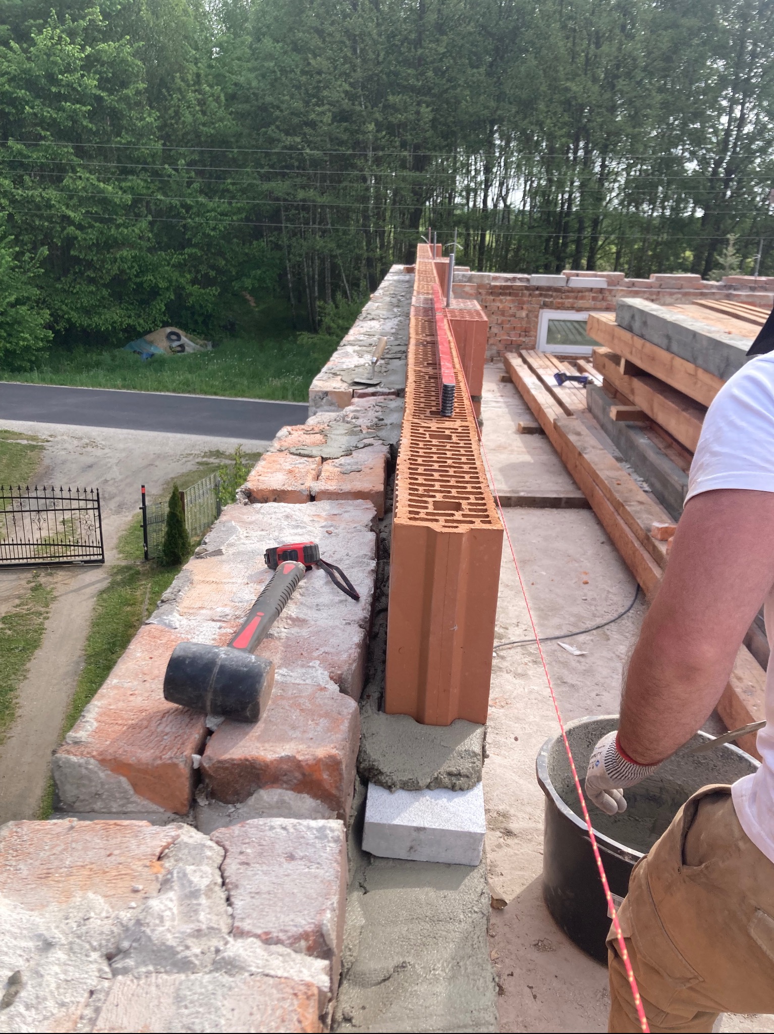 Murowanie ściany z pustaków ceramicznych Porotherm. Widoczny murarz z kielnią i zaprawą, poziomica, sznurek murarski. W tle drzewa i droga.