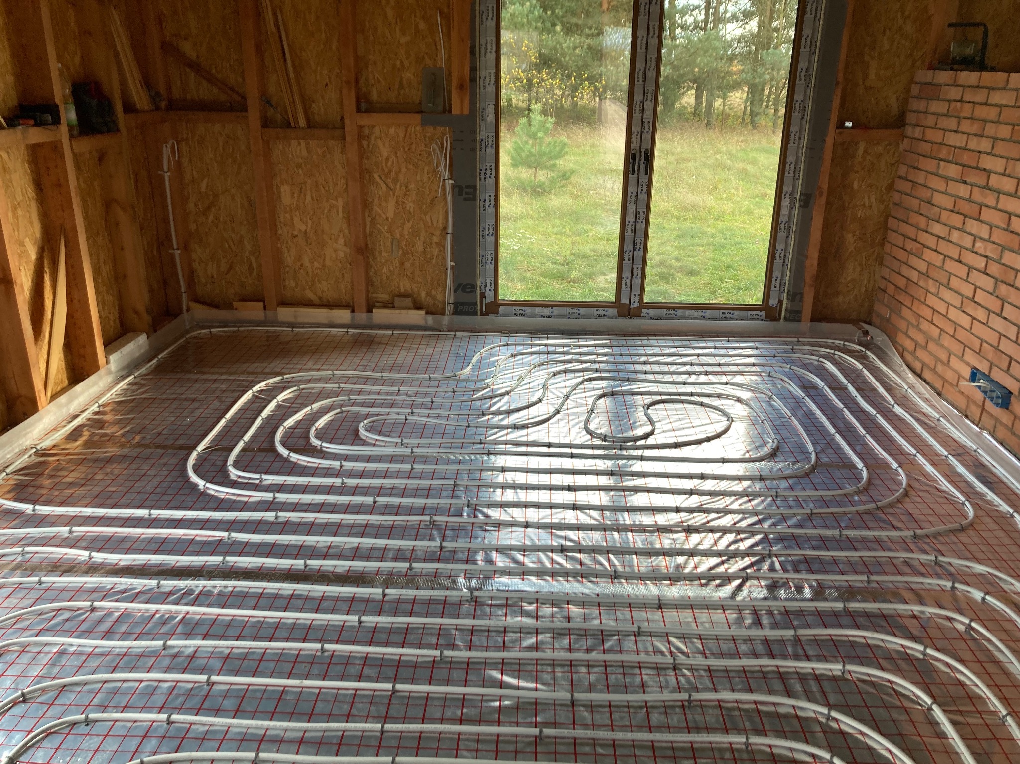 Instalacja ogrzewania podłogowego w surowym wnętrzu z oknem na zewnątrz. Widoczne rury na folii z siatką, ściany z płyty OSB i cegły. Etap prac przed wylaniem posadzki.