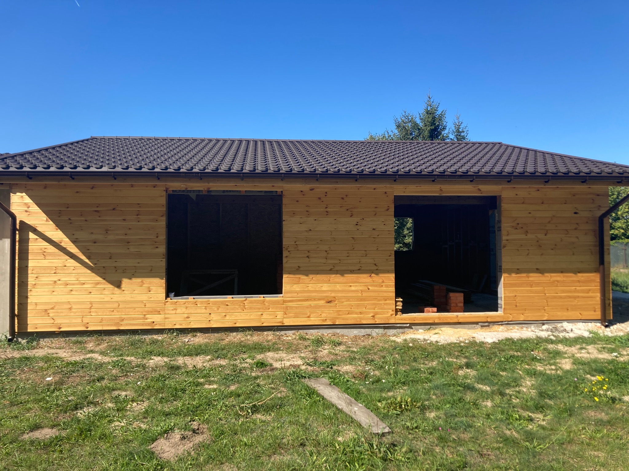 Drewniany budynek w trakcie budowy z brązowym dachem na tle błękitnego nieba. Widoczne niezabudowane otwory okienne i drzwiowe. Budynek otoczony zielenią.