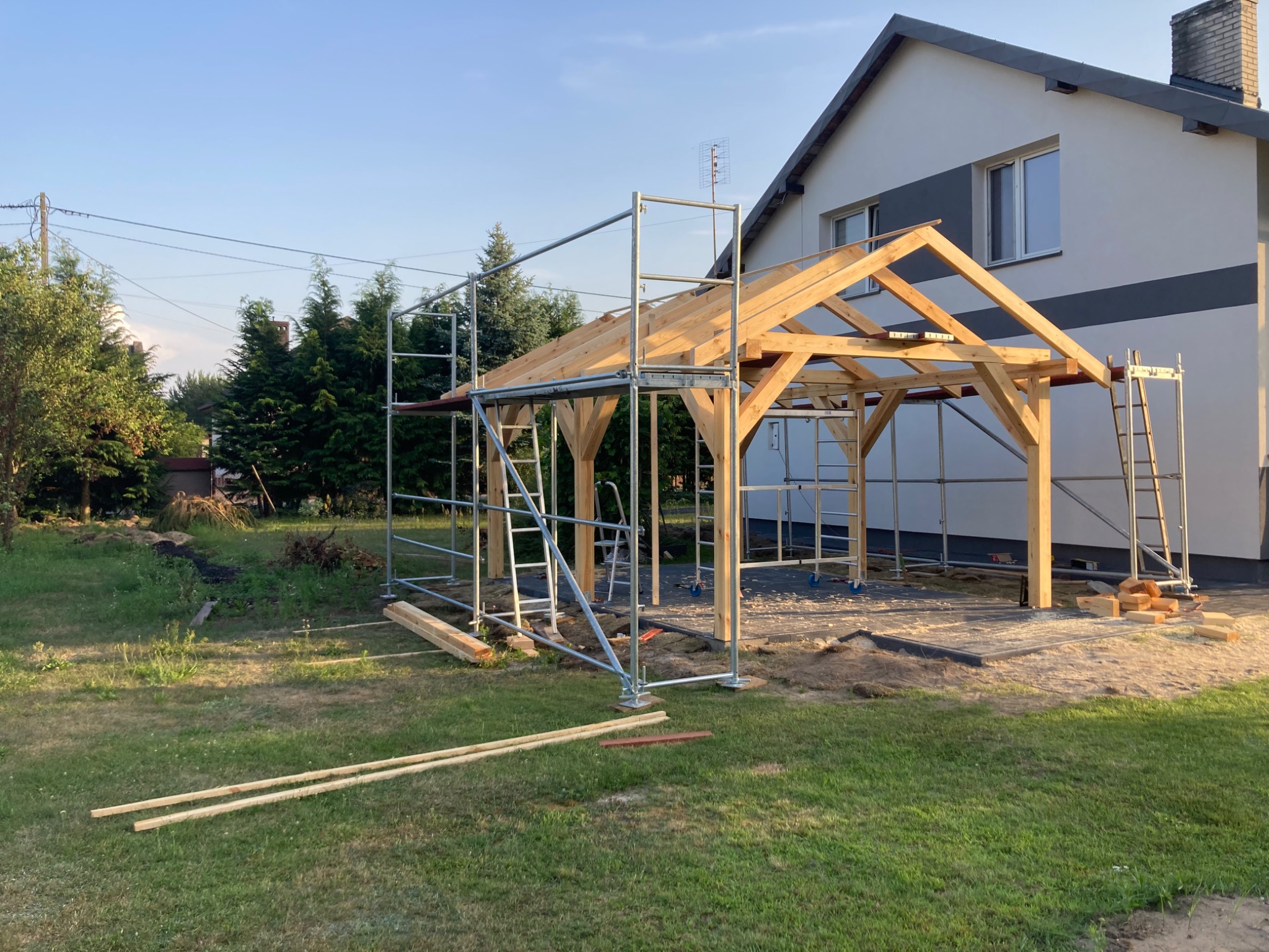 Drewniana konstrukcja zadaszenia w trakcie budowy, otoczona rusztowaniami na trawiastym terenie, z domem w tle. Widoczne elementy konstrukcyjne i narzędzia.