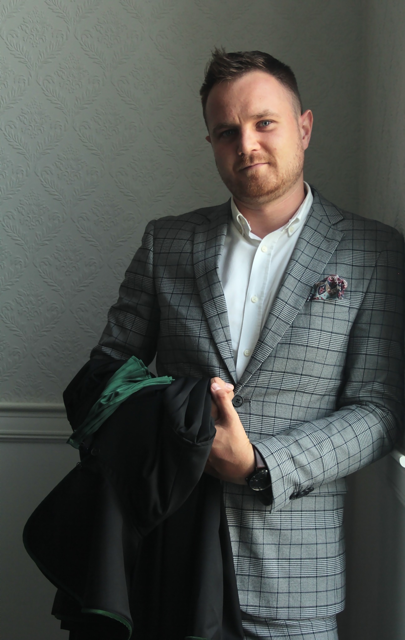 Portret młodego adwokata w kraciastej marynarce, trzymającego togę z zielonym wykończeniem, opierającego się o ścianę z delikatnym wzorem.