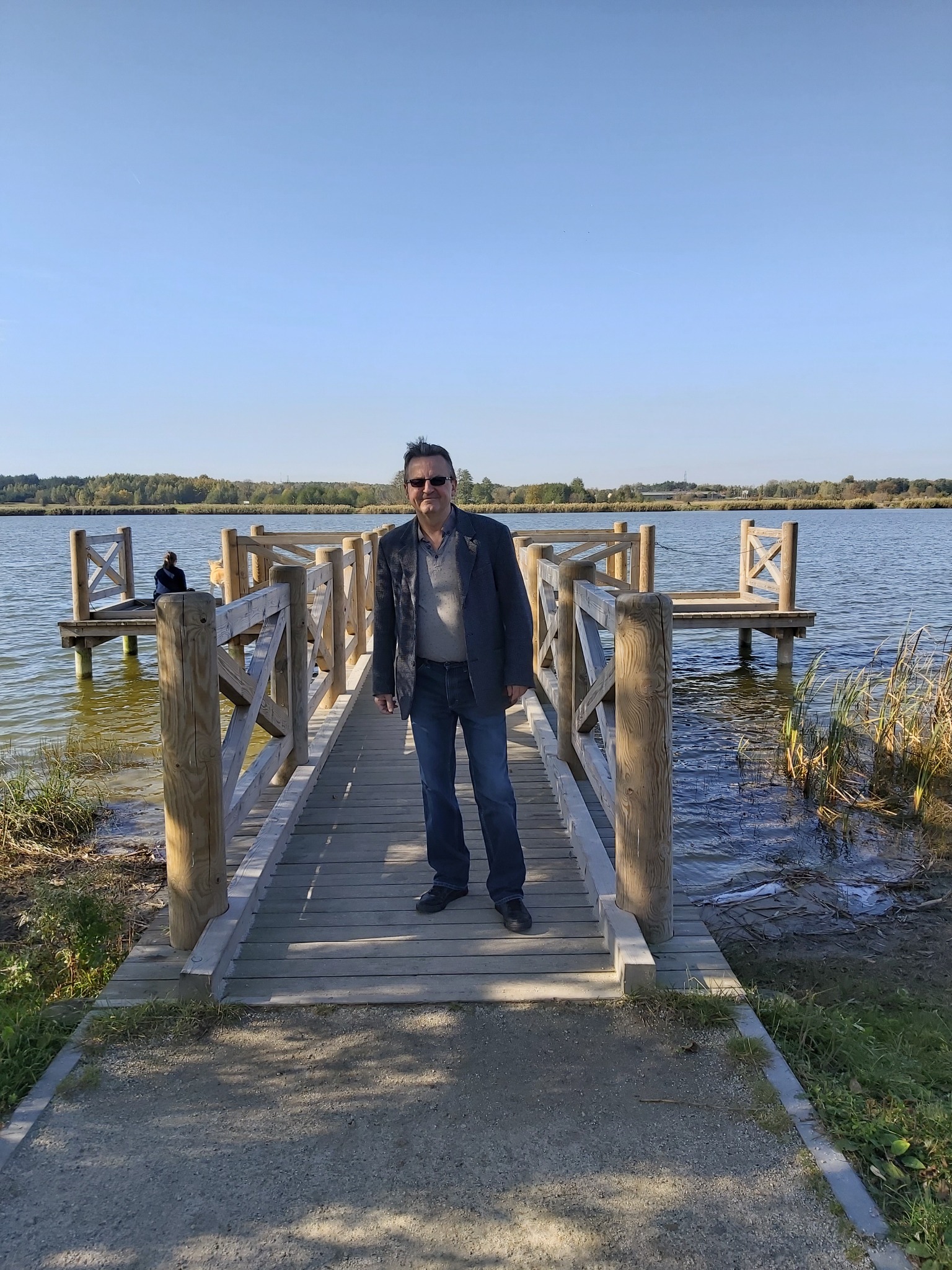 Mężczyzna w okularach i kurtce stoi na drewnianym pomoście nad jeziorem w słoneczny dzień. W tle widoczny las i błękitne niebo.