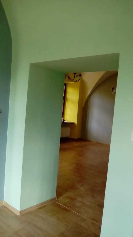 Pomalowane na jasnozielono ściany i futryna drzwiowa, widok na sąsiednie pomieszczenie z żółtą ścianą i drewnianą podłogą. Widoczny fragment żyrandola.