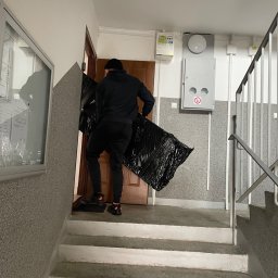 Przewózka - Pracownik w czarnej odzieży wnosi zabezpieczony folią mebel po schodach klatki schodowej do mieszkania. Widoczne schody, ściany i fragment drzwi.