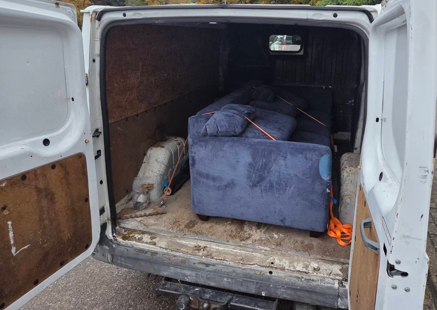 Wnętrze białego busa z otwartymi drzwiami, w którym znajduje się zabezpieczona pomarańczowymi pasami niebieska sofa z poduszkami. Widoczny hak holowniczy.