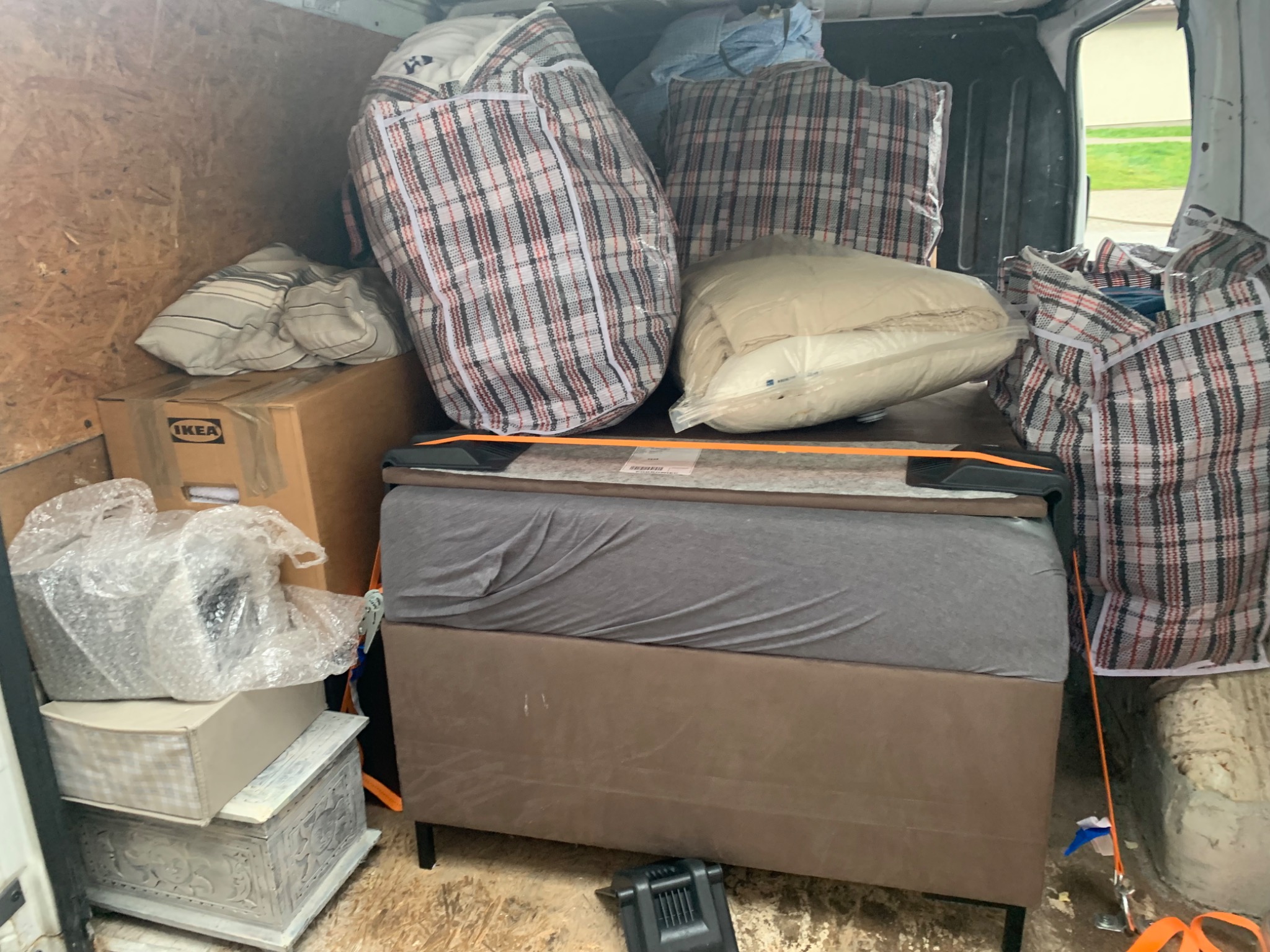 Wnętrze furgonetki załadowane workami w kratę, kartonem IKEA i meblami, zabezpieczone pomarańczowym pasem. Widoczne ślady użytkowania na podłodze i ścianach.