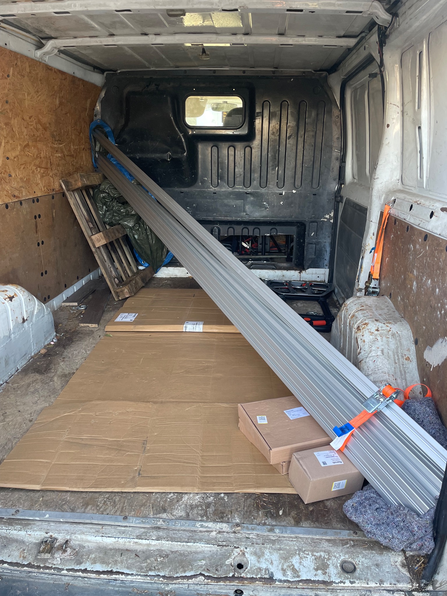 Wnętrze brudnego busa z zabezpieczonym ładunkiem: długie profile aluminiowe, kartony i paleta. Widoczne pasy mocujące i kartonowe podkładki na podłodze.