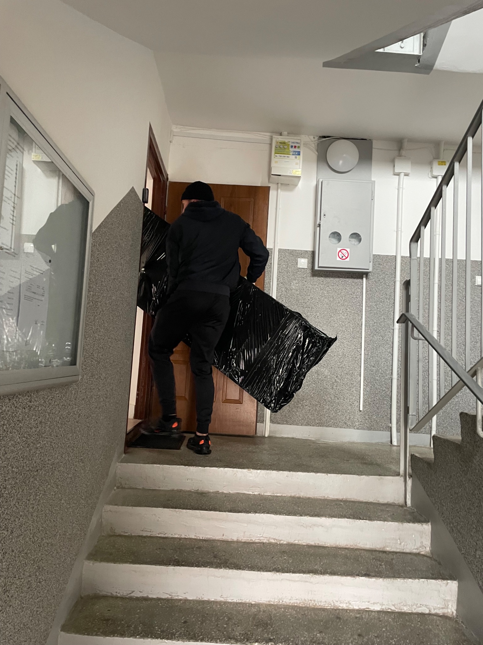 Pracownik w czarnej odzieży wnosi zabezpieczony folią mebel po schodach klatki schodowej do mieszkania. Widoczne schody, ściany i fragment drzwi.