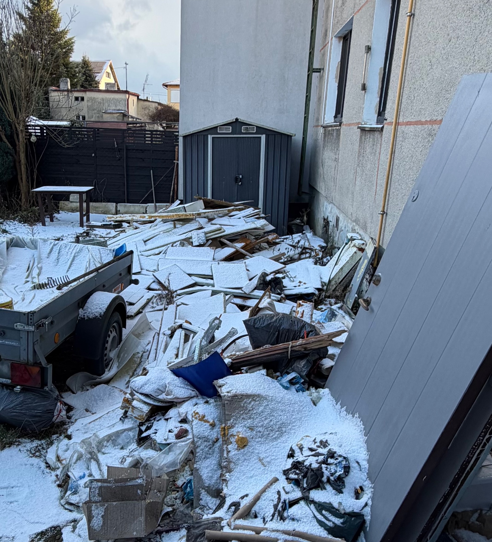 Zasypany śniegiem teren po opróżnianiu, z przyczepą i stertą gruzu przy budynku. Widoczny garaż blaszany i elementy wyposażenia. Zimowy krajobraz po demontażu.